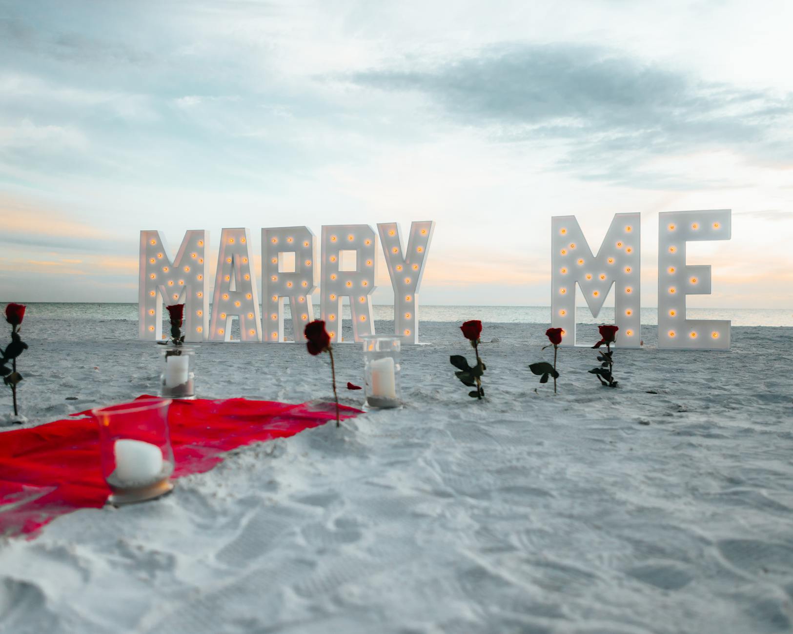 Romantyczne zaręczyny na plaży z różami i podświetlanym napisem Marry Me